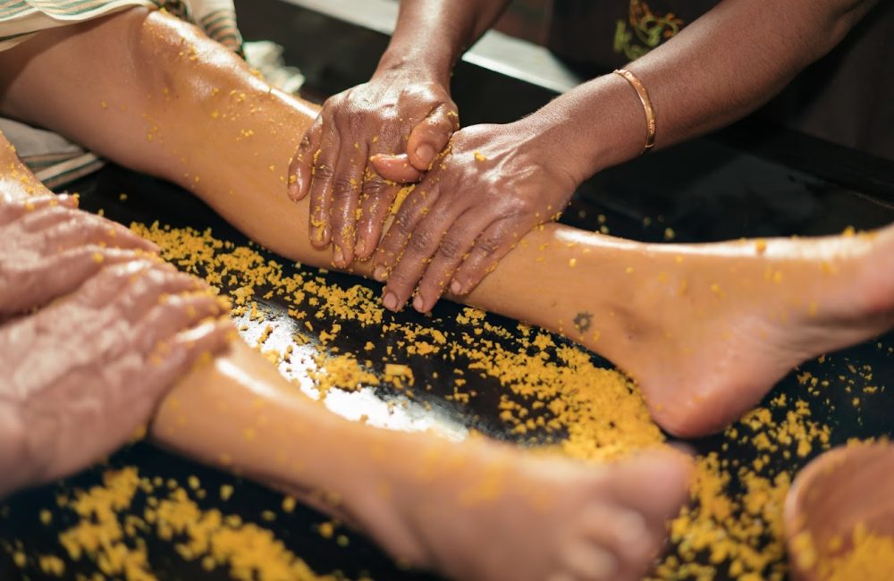 Hands applying golden turmeric paste or scrub to legs and feet during a spa treatment or traditional wellness ritual
