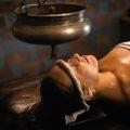 Woman receiving traditional Ayurvedic Shirodhara therapy with warm herbal oil gently poured on the forehead.