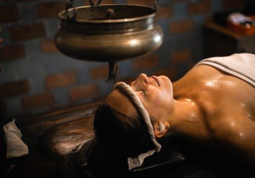 Woman receiving traditional Ayurvedic Shirodhara therapy with warm herbal oil gently poured on the forehead.