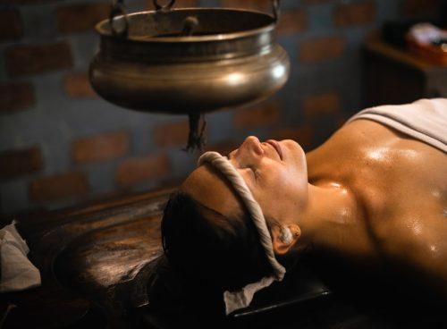 Woman receiving traditional Ayurvedic Shirodhara therapy with warm herbal oil gently poured on the forehead.