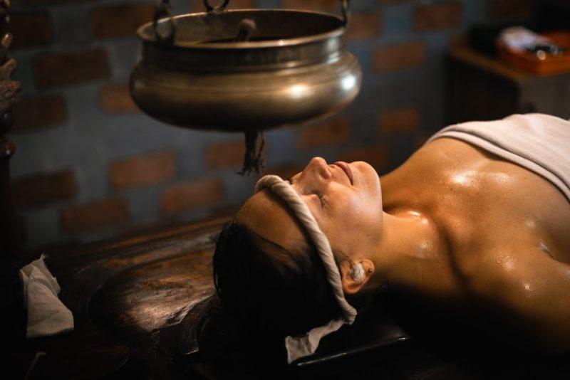 Woman receiving traditional Ayurvedic Shirodhara therapy with warm herbal oil gently poured on the forehead.