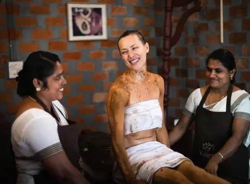 Smiling woman receiving Ayurvedic herbal therapy at Kairali The Ayurvedic Healing Village in Kerala, assisted by two female therapists in traditional attire.