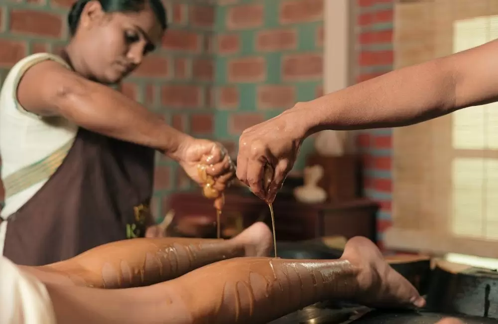 Two Ayurvedic therapists performing traditional oil therapy on a patient’s legs using warm herbal oils at Kairali Ayurvedic Health Retreat in Palakkad, Kerala.