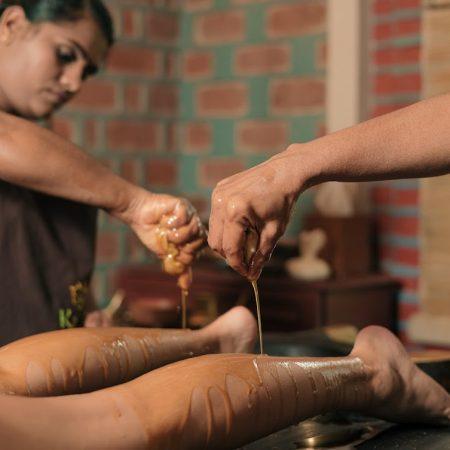 Two Ayurvedic therapists performing traditional oil therapy on a patient’s legs using warm herbal oils at Kairali Ayurvedic Health Retreat in Palakkad, Kerala.