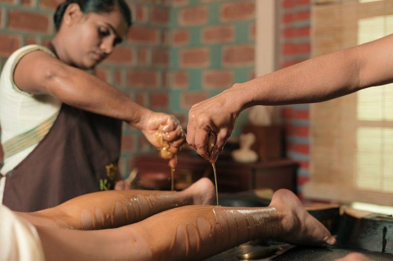 Two Ayurvedic therapists performing traditional oil therapy on a patient’s legs using warm herbal oils at Kairali Ayurvedic Health Retreat in Palakkad, Kerala.