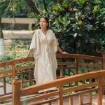 Woman in Ayurvedic robe standing on a wooden bridge at Kairali Ayurvedic Health Village in Kerala, surrounded by lush greenery.