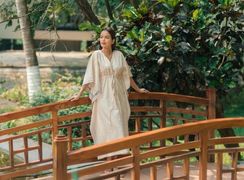 Woman in Ayurvedic robe standing on a wooden bridge at Kairali Ayurvedic Health Village in Kerala, surrounded by lush greenery.