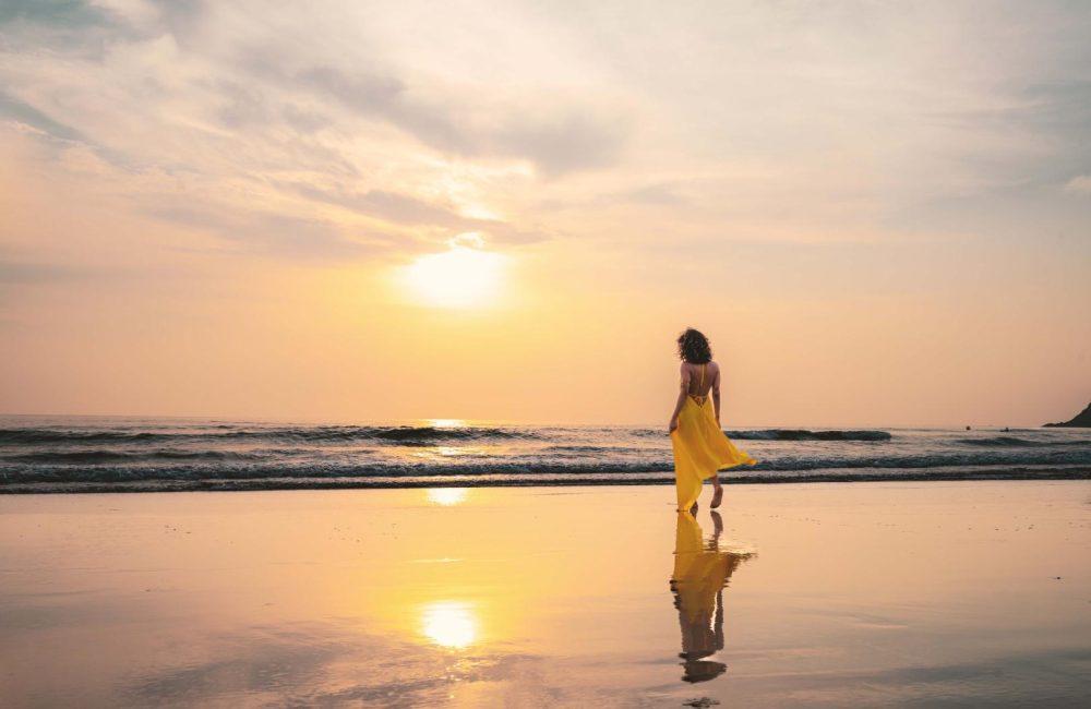 Woman in a flowing yellow dress walking along Agonda Beach at sunset, soft waves and mirrored reflection on wet sand, South Goa.