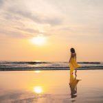 Woman in a flowing yellow dress walking along Agonda Beach at sunset, soft waves and mirrored reflection on wet sand, South Goa.