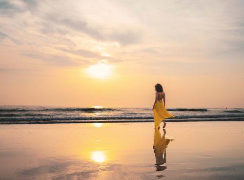 Woman in a flowing yellow dress walking along Agonda Beach at sunset, soft waves and mirrored reflection on wet sand, South Goa.