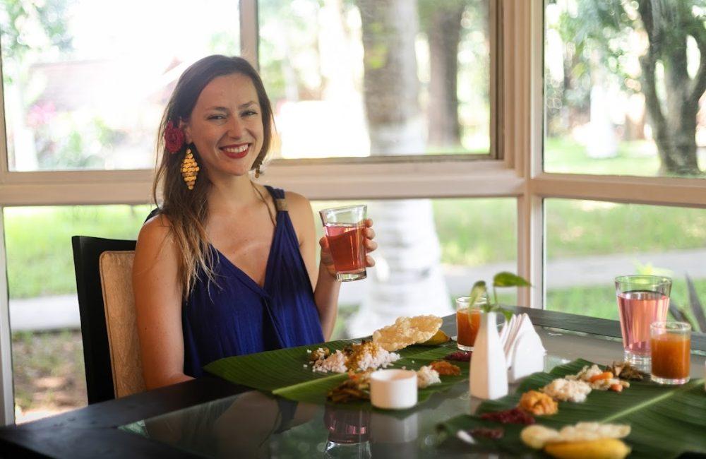 Smiling solo guest holding a pink herbal drink while enjoying a banana-leaf Ayurvedic meal in a garden-view dining pavilion at Kairali Ayurvedic Healing Village, Kerala.