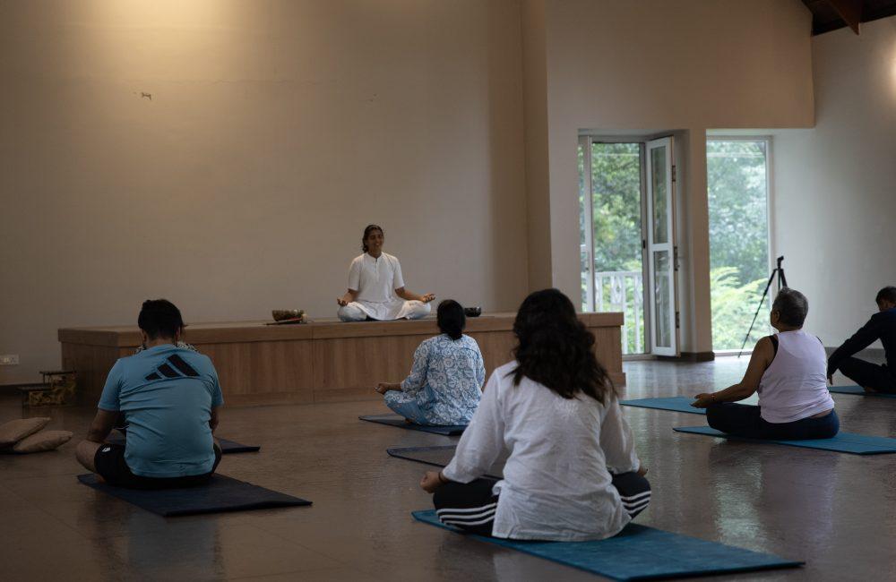 Group meditation class seated on mats facing a teacher on a low stage in a serene hall with open doors and greenery outside at a Kerala yoga retreat.
