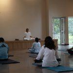 Group meditation class seated on mats facing a teacher on a low stage in a serene hall with open doors and greenery outside at a Kerala yoga retreat.