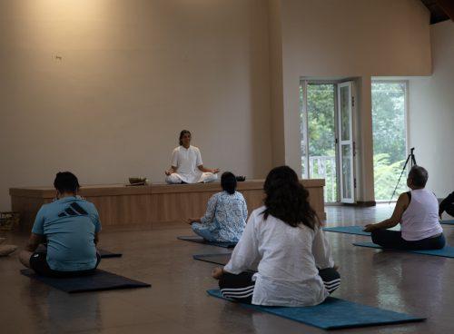 Group meditation class seated on mats facing a teacher on a low stage in a serene hall with open doors and greenery outside at a Kerala yoga retreat.
