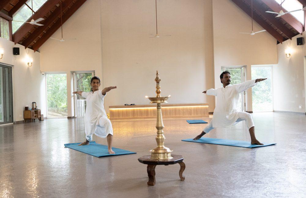 Two practitioners in white perform Virabhadrasana II on blue mats beside a lit brass nilavilakku in a serene hall at a yoga retreat in Kerala.