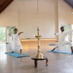 Two practitioners in white perform Virabhadrasana II on blue mats beside a lit brass nilavilakku in a serene hall at a yoga retreat in Kerala.