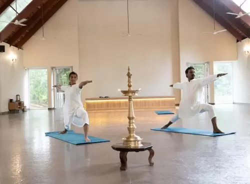 Two practitioners in white perform Virabhadrasana II on blue mats beside a lit brass nilavilakku in a serene hall at a yoga retreat in Kerala.