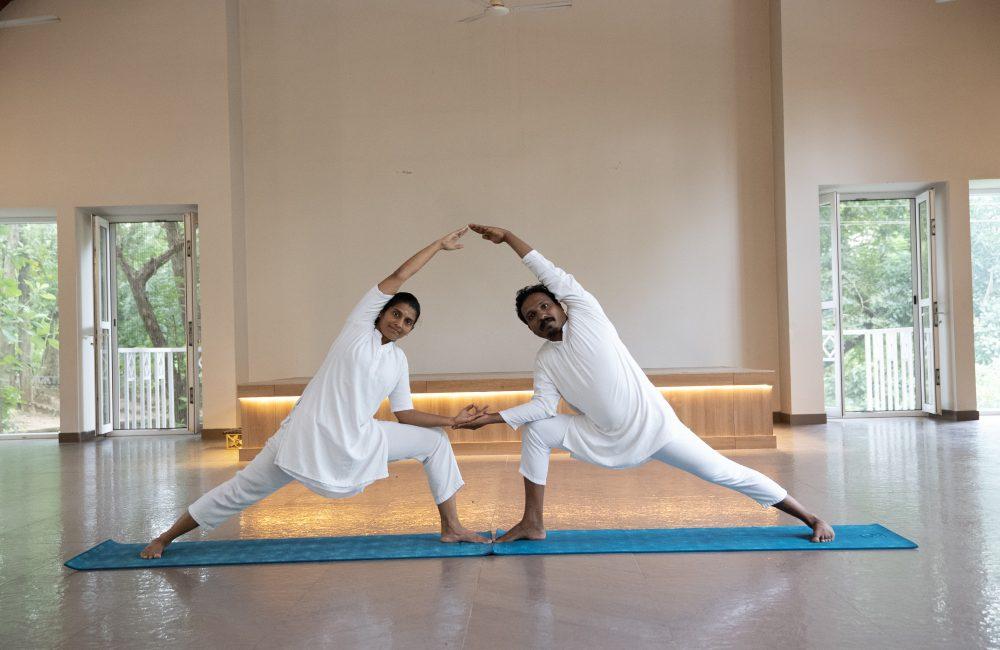 Two yoga practitioners in mirrored side-angle pose on a blue mat inside kairali - The Ayurvedic Healing Village with open doors and greenery outside.