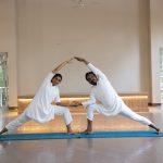 Two yoga practitioners in mirrored side-angle pose on a blue mat inside kairali - The Ayurvedic Healing Village with open doors and greenery outside.