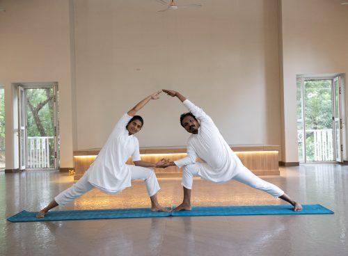 Two yoga practitioners in mirrored side-angle pose on a blue mat inside kairali - The Ayurvedic Healing Village with open doors and greenery outside.