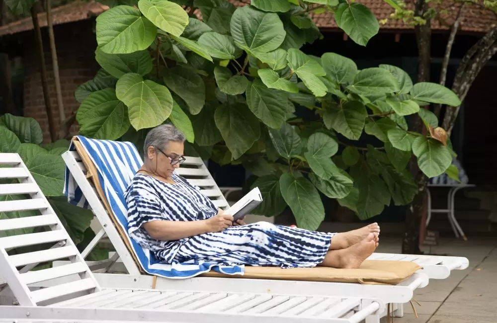 Senior woman relaxing on a lounge chair, reading under lush green leaves at Kairali Ayurvedic Healing Village in Kerala.