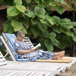 Senior woman relaxing on a lounge chair, reading under lush green leaves at Kairali Ayurvedic Healing Village in Kerala.