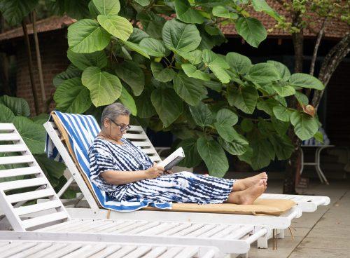 Senior woman relaxing on a lounge chair, reading under lush green leaves at Kairali Ayurvedic Healing Village in Kerala.