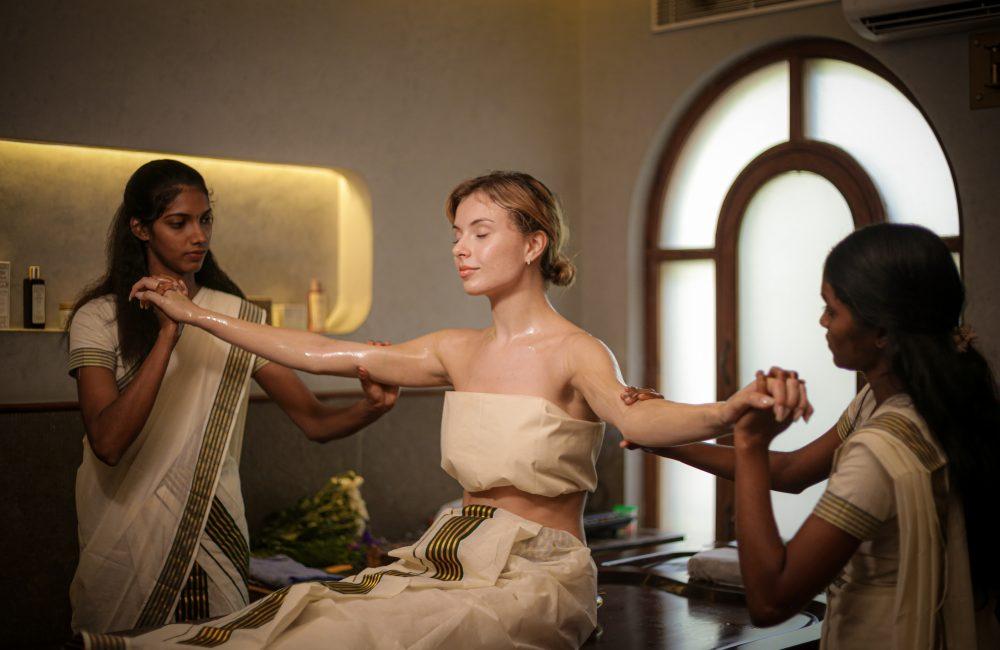 Woman receiving traditional Ayurvedic abhyanga oil therapy from two female therapists in a serene Kerala retreat centre.