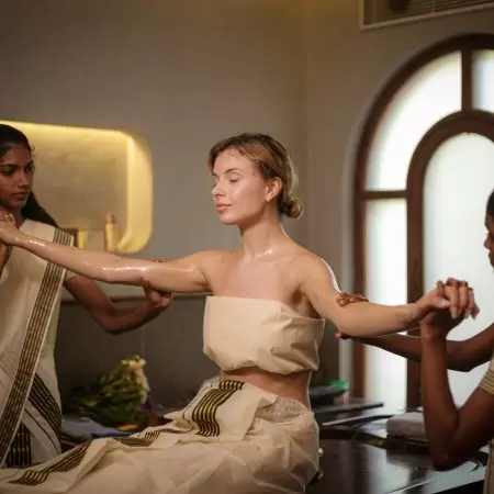 Woman receiving traditional Ayurvedic abhyanga oil therapy from two female therapists in a serene Kerala retreat centre.