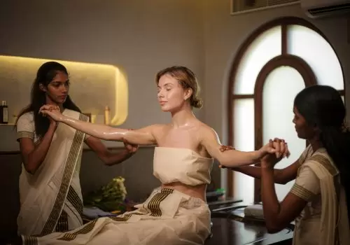 Woman receiving traditional Ayurvedic abhyanga oil therapy from two female therapists in a serene Kerala retreat centre.