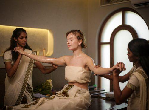 Woman receiving traditional Ayurvedic abhyanga oil therapy from two female therapists in a serene Kerala retreat centre.