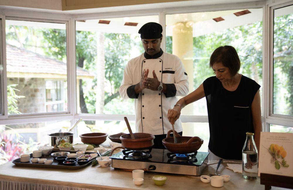Chef leading an Ayurvedic cooking class at Kairali – The Ayurvedic Healing Village, guiding a guest as clay pots simmer beside spice trays and cookbooks.