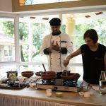 Chef leading an Ayurvedic cooking class at Kairali – The Ayurvedic Healing Village, guiding a guest as clay pots simmer beside spice trays and cookbooks.