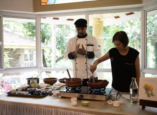 Chef leading an Ayurvedic cooking class at Kairali – The Ayurvedic Healing Village, guiding a guest as clay pots simmer beside spice trays and cookbooks.