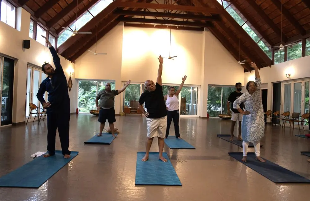 Senior guests practicing gentle side-bend yoga on mats in a spacious, well-lit hall at Kairali Ayurvedic Health Village Kerala—an accessible ayurvedic retreat Kerala.