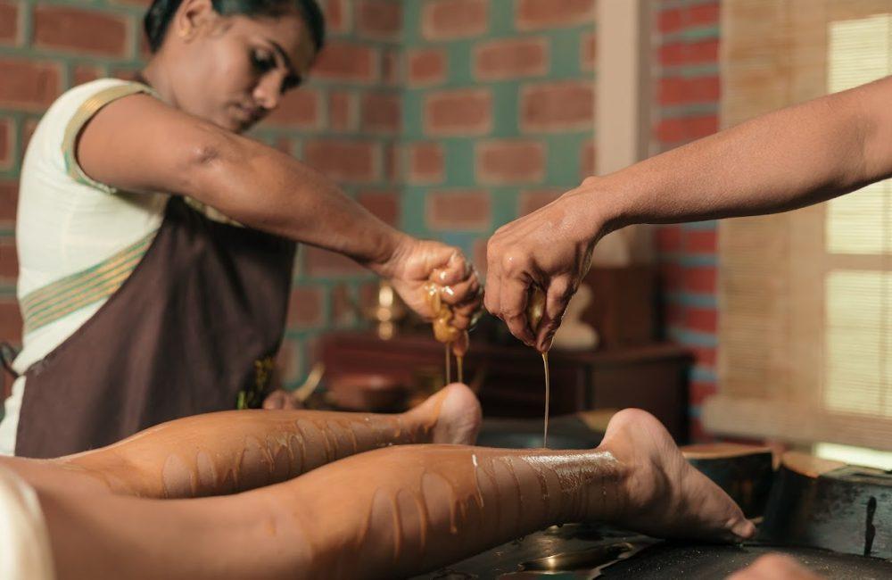 Therapists pour warm herbal oil onto a guest’s legs during adapted Ayurveda care at Kairali—The Ayurvedic Healing Village, Kerala.