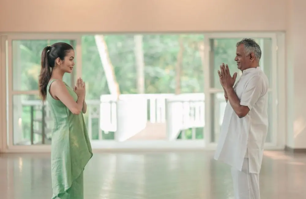 Two people greet in Namaste inside a sunlit yoga studio at Kairali—The Ayurvedic Healing Village, Palakkad, Kerala, surrounded by lush greenery.