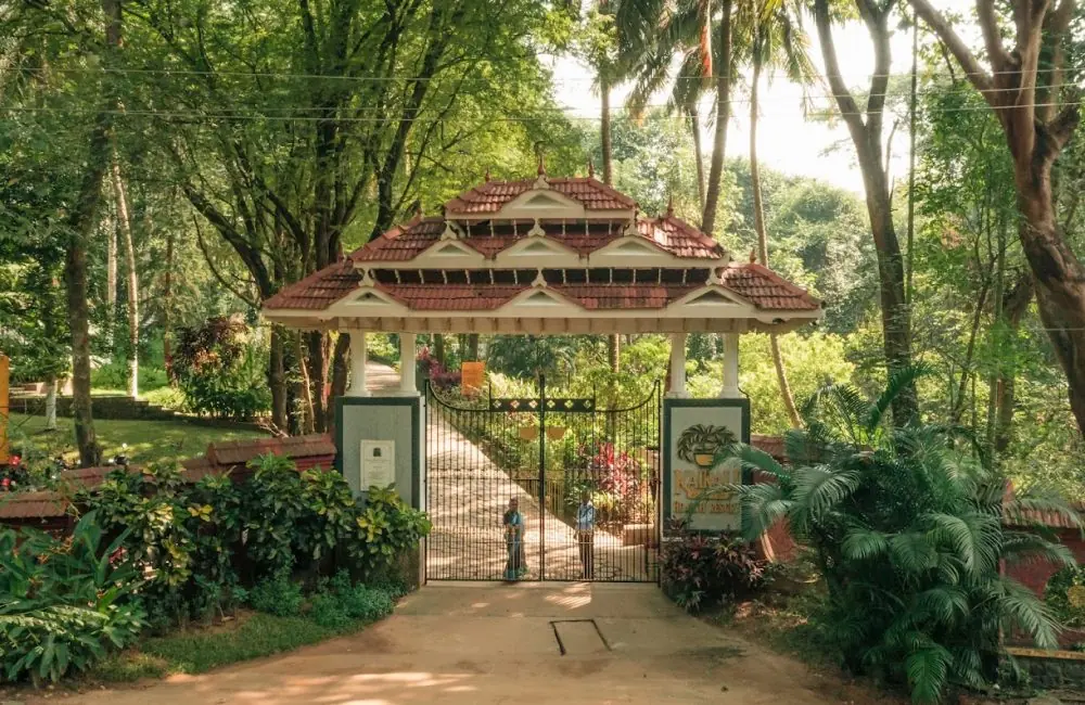 Kerala-style tiled entrance gate of Kairali—The Ayurvedic Healing Village in Palakkad, framed by lush trees, with a pathway and sign visible.