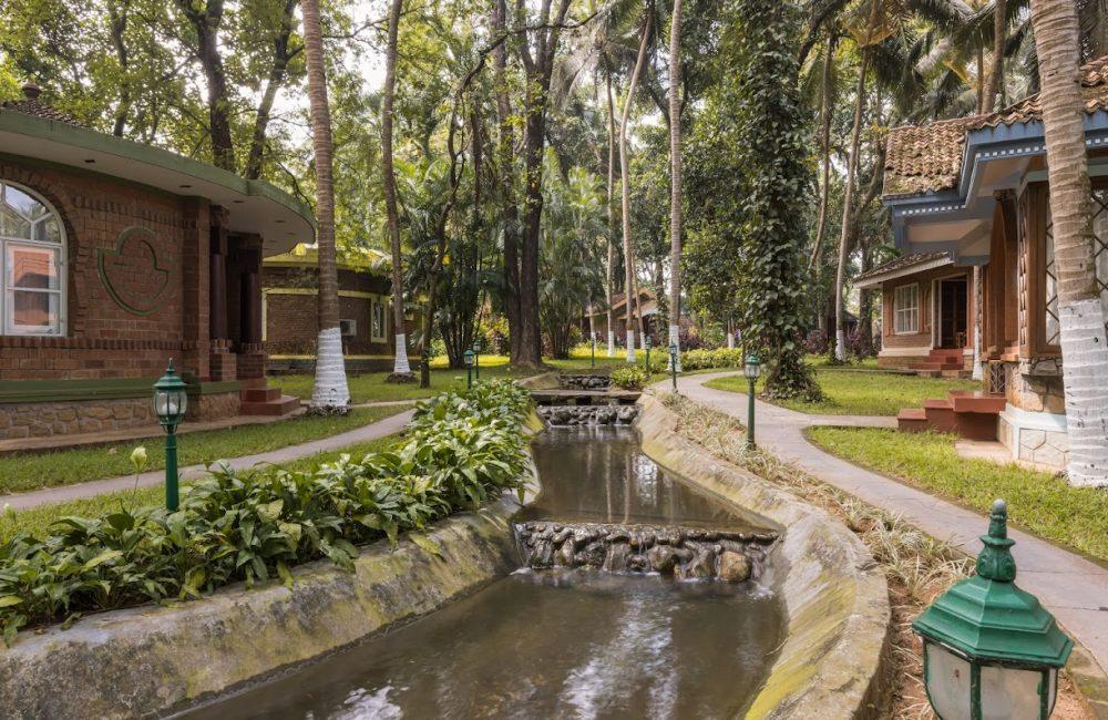 Tree-shaded garden stream with red-brick cottages and walking paths at Kairali retreat Kerala—quiet Ayurvedic campus ideal for privacy-first stays.