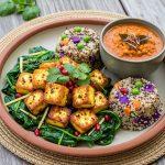 Golden pan-seared paneer on sautéed spinach with mustard seeds, quinoa-millet vegetable mounds, and a bowl of red lentil dal; garnished with cilantro and pomegranate on a terracotta plate.