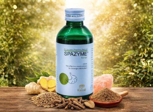 Green bottle of Kairali Spazyme Syrup (200 ml) placed outdoors on a wooden surface, surrounded by fennel seeds, long pepper, ginger, rock salt, and a bowl of ground herbs, representing natural digestive care.