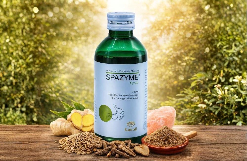 Green bottle of Kairali Spazyme Syrup (200 ml) placed outdoors on a wooden surface, surrounded by fennel seeds, long pepper, ginger, rock salt, and a bowl of ground herbs, representing natural digestive care.