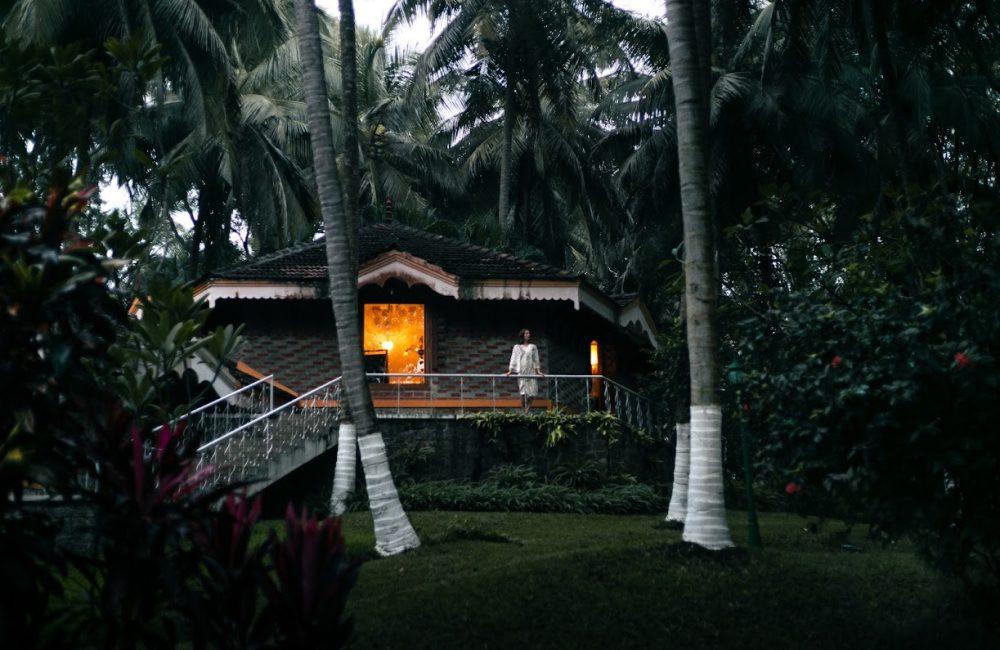 Solo woman on the veranda of a warmly lit Kerala cottage at Kairali – The Ayurvedic Healing Village, surrounded by coconut palms at dusk.