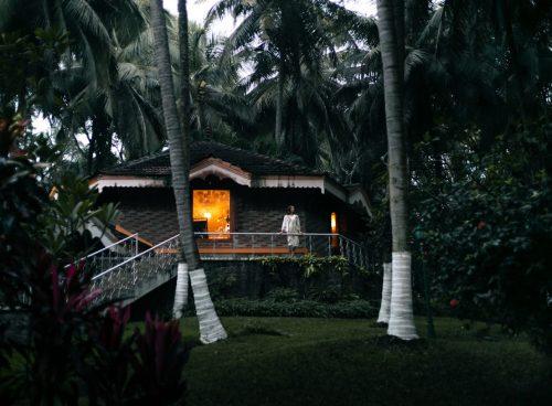 Solo woman on the veranda of a warmly lit Kerala cottage at Kairali – The Ayurvedic Healing Village, surrounded by coconut palms at dusk.