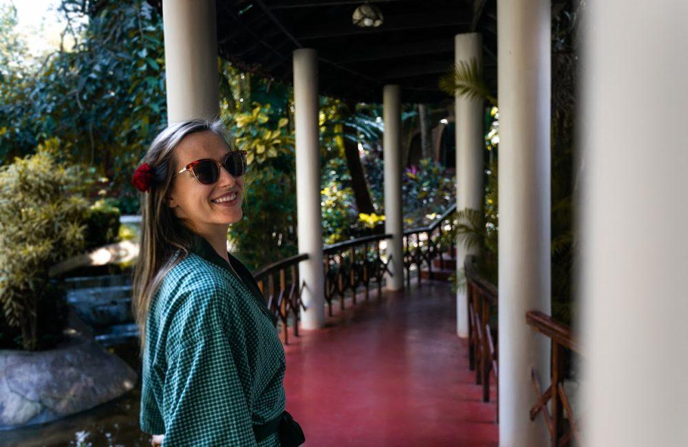 Smiling solo woman in a green robe with a flower in her hair walking along a shaded, columned walkway at Kairali – The Ayurvedic Healing Village, Kerala.