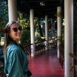 Smiling solo woman in a green robe with a flower in her hair walking along a shaded, columned walkway at Kairali – The Ayurvedic Healing Village, Kerala.