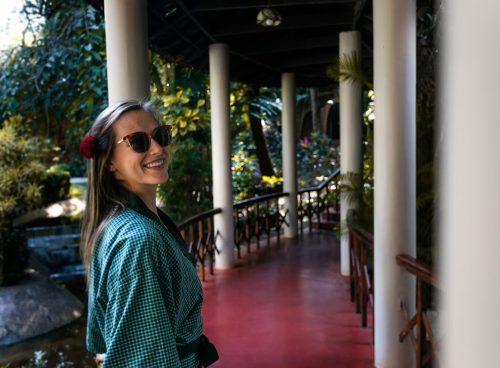 Smiling solo woman in a green robe with a flower in her hair walking along a shaded, columned walkway at Kairali – The Ayurvedic Healing Village, Kerala.