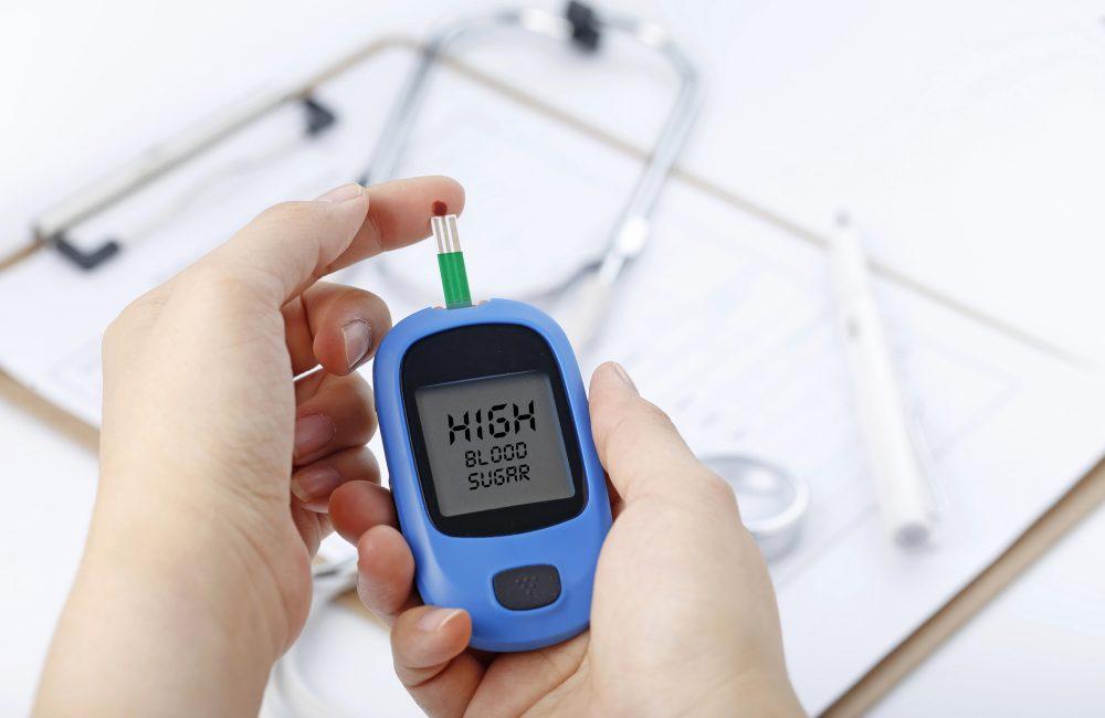 Hands holding a glucometer reading “HIGH BLOOD SUGAR,” with a blood drop on the test strip; stethoscope and chart blurred in the background.