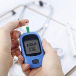 Hands holding a glucometer reading “HIGH BLOOD SUGAR,” with a blood drop on the test strip; stethoscope and chart blurred in the background.