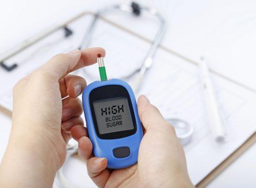 Hands holding a glucometer reading “HIGH BLOOD SUGAR,” with a blood drop on the test strip; stethoscope and chart blurred in the background.